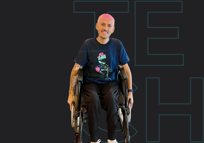 Leandro esta sorrindo e sentado numa cadeira de rodas. Ele tem o cabelo rosa curto, tatuagens no braço e usa uma blusa azul com um desenho na parte central e calça preta.